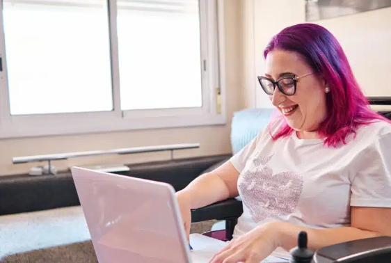 Image of a woman in a wheelchair with pink hair seated at her computer