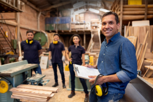 Three people in hard hats and a man in forground wearing blue uniform, smiling to camera in a wood workshop