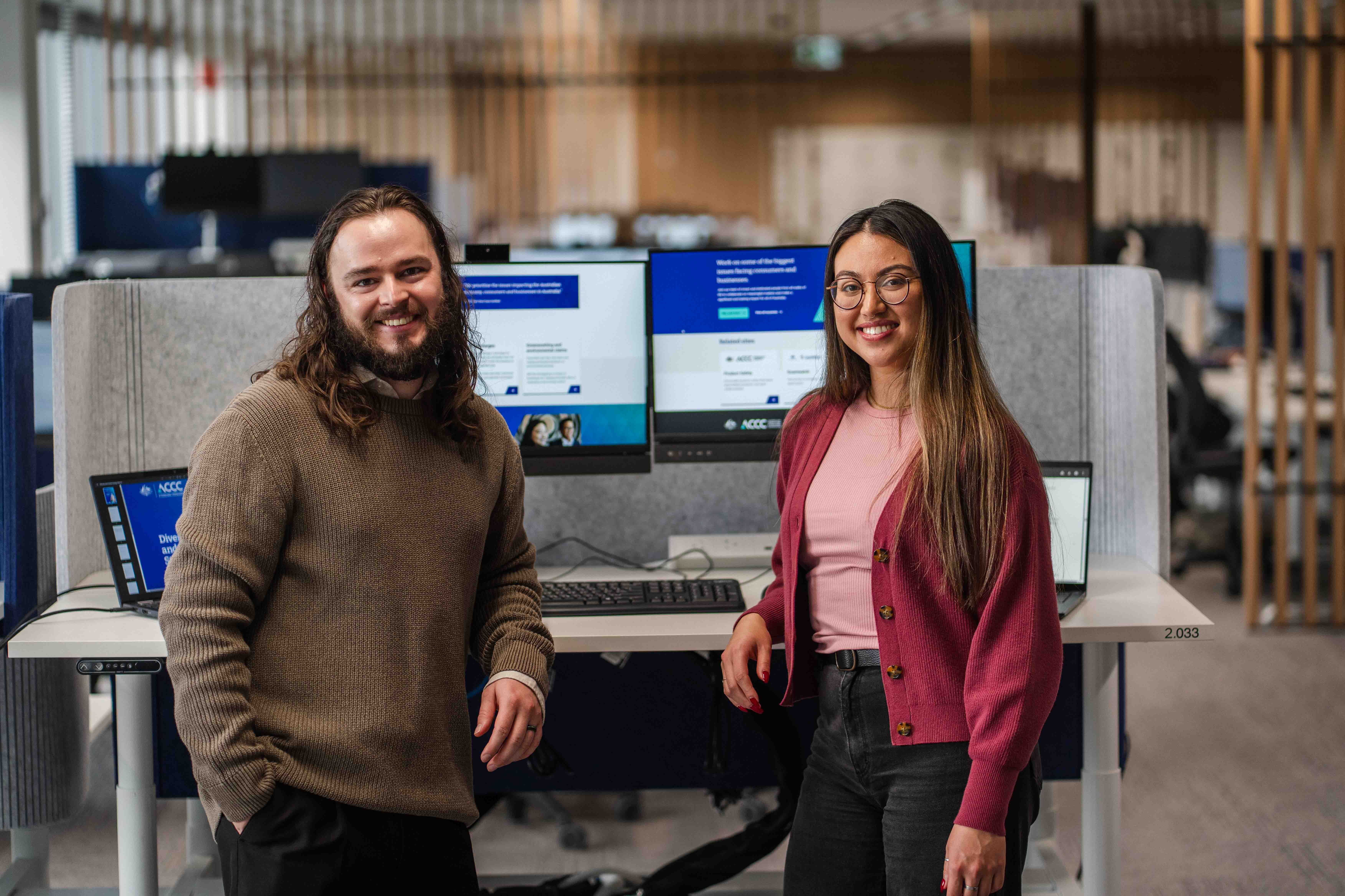 Photo from ACCC with a man and a woman on either side of a computer, looking at the camera and smiling