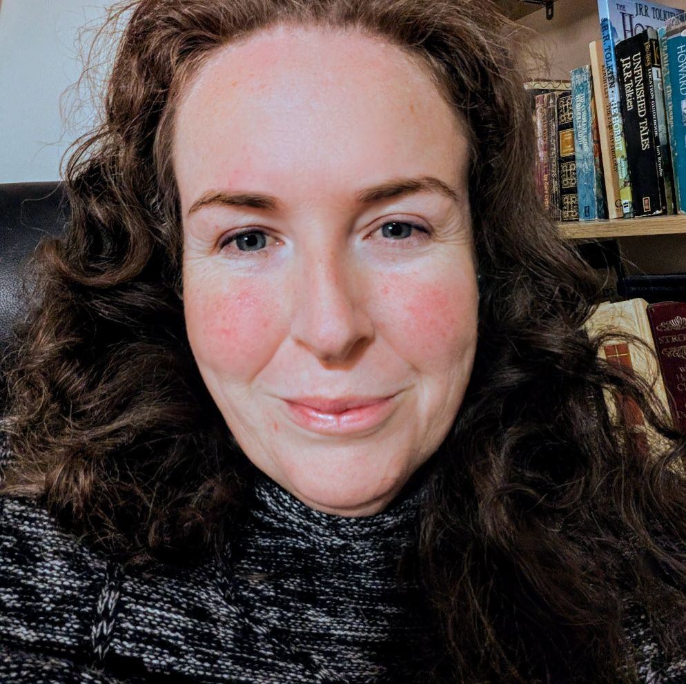Headshot of Alisha Taylor-Jones who is a white woman with brown, curly hair. She wears a high-neck jumper and sits in front of a bookshelf.