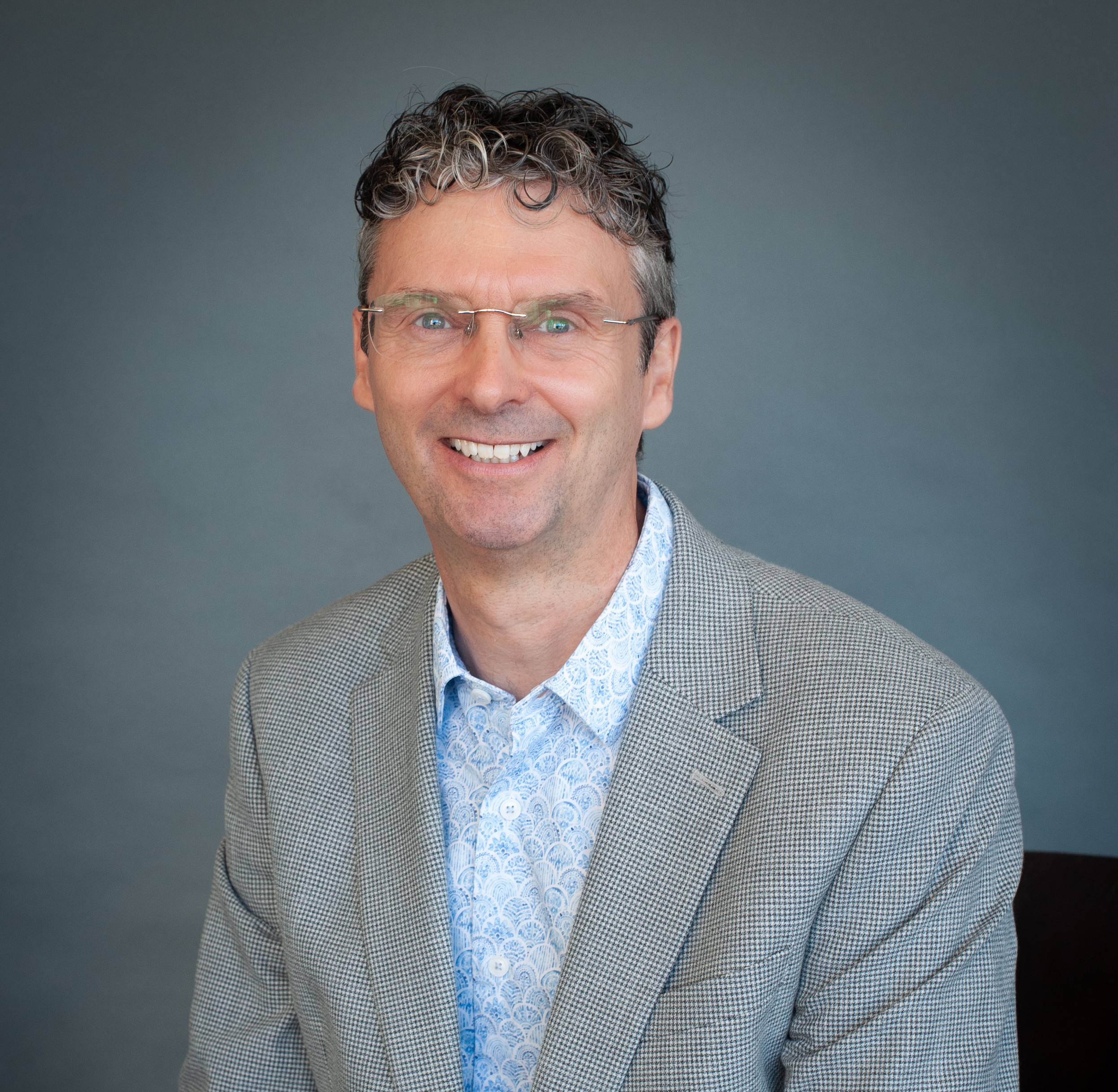 Headshot of Andy Brown, a white man with curly, grey hair, glasses and a grey and light blue suit.