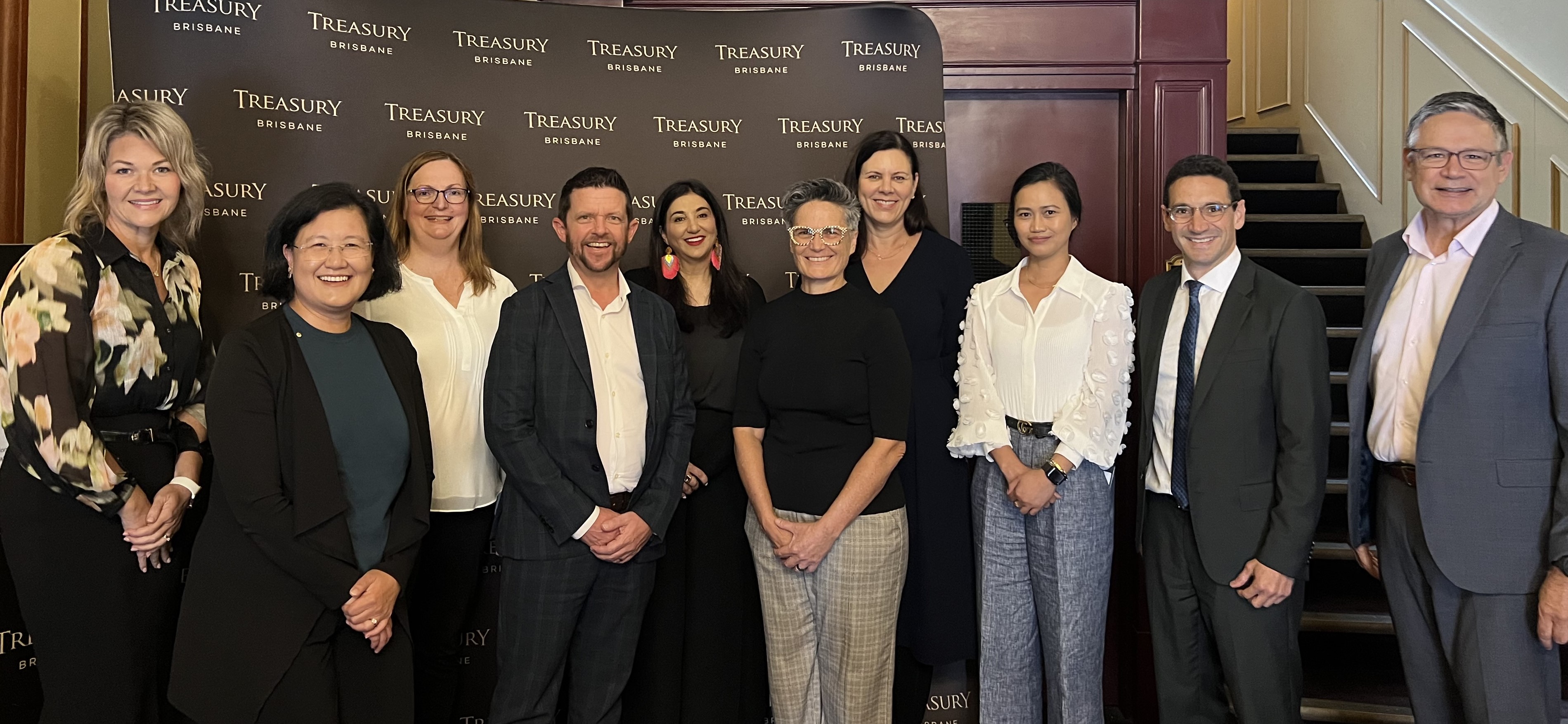Company and organisation leaders pose for a photo in front of a banner with the words Treasury Brisbane on it. The people are wearing business attire and smiling.
