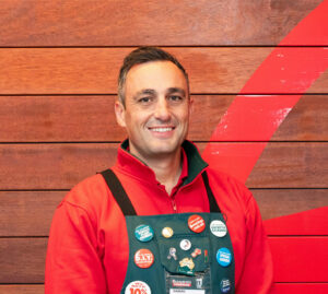 Damian Zahra appears in front of a wood panelled wall with the Bunnings logo, wearing a red  and green Bunnings uniform.