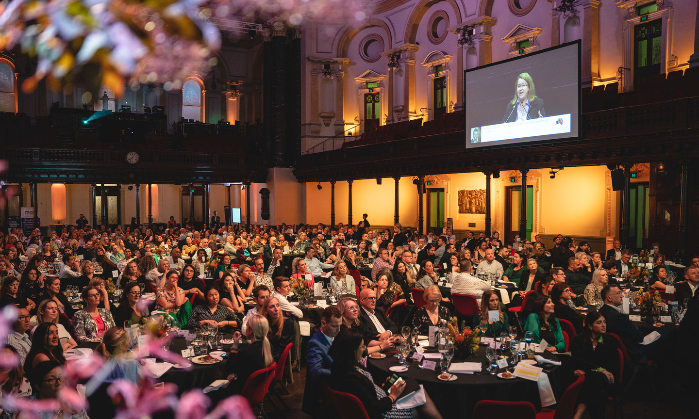 Audience members in formal clothing sit around dining tables in a lit up hall, looking at speaker on stage.