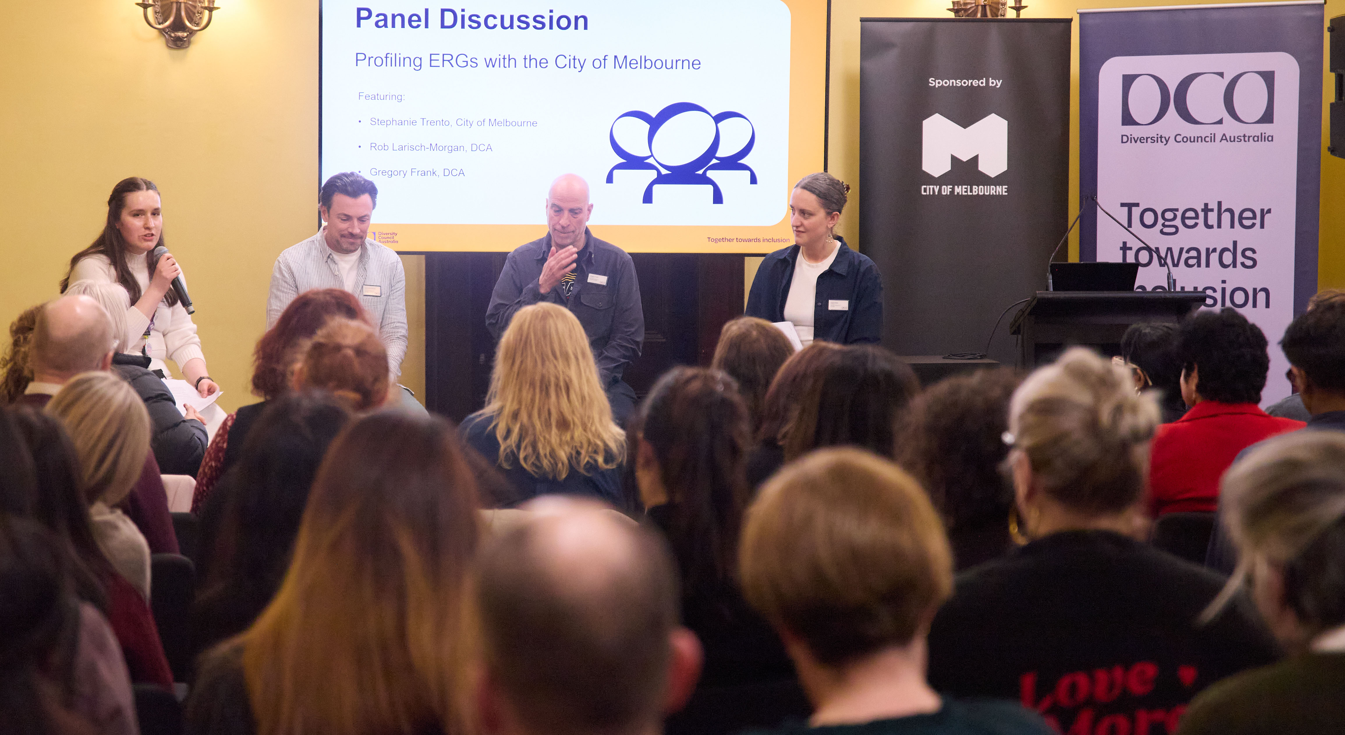 Shot from behind of DCA members looking at stage with four people presenting on a panel at the Key Contact even in Melbourne