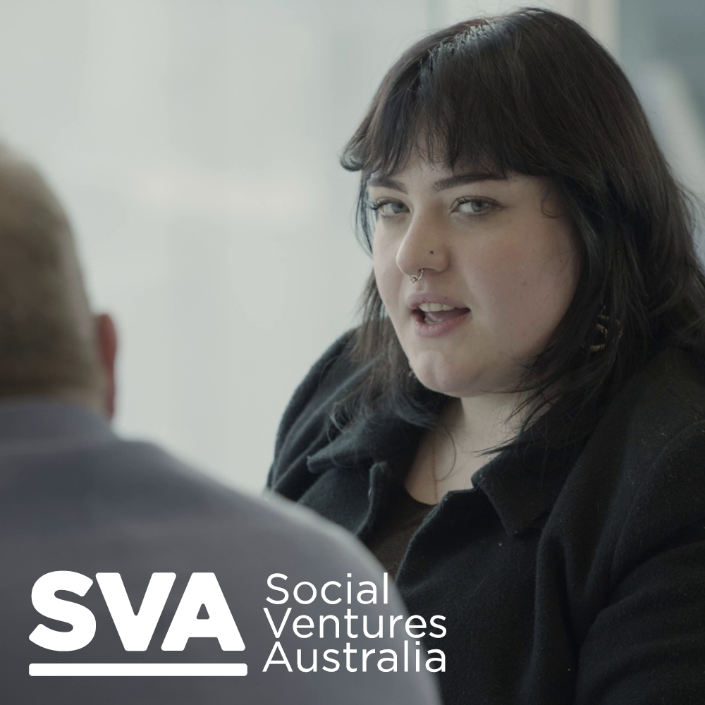 Social Ventures Australia logo in white, in the background a white woman with dark hair and black clothes, speaking while looking at the camera
