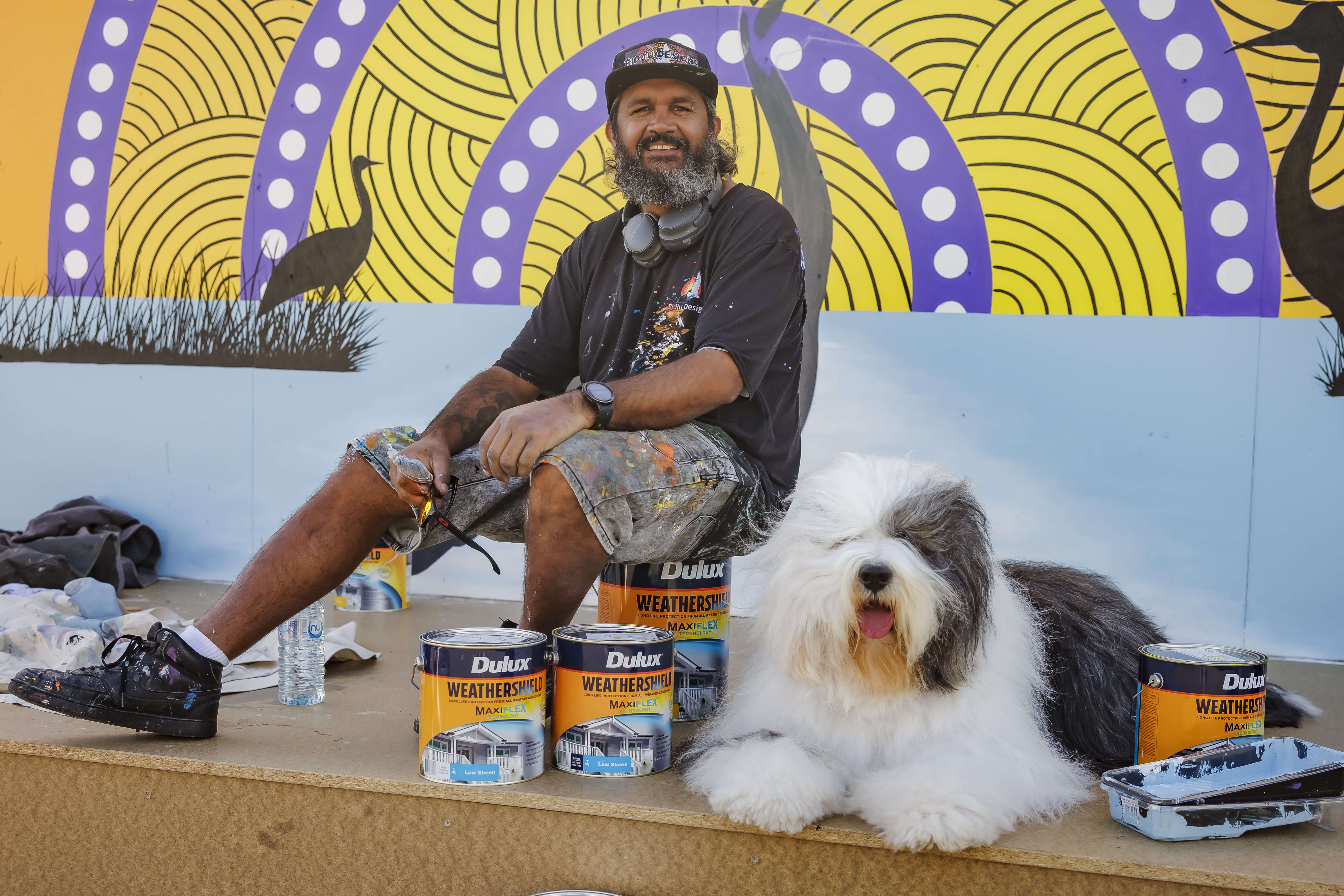Aboriginal man sits on a bucket of Dulux paint next to a fluffy dog and behind a yellow and purple mural.