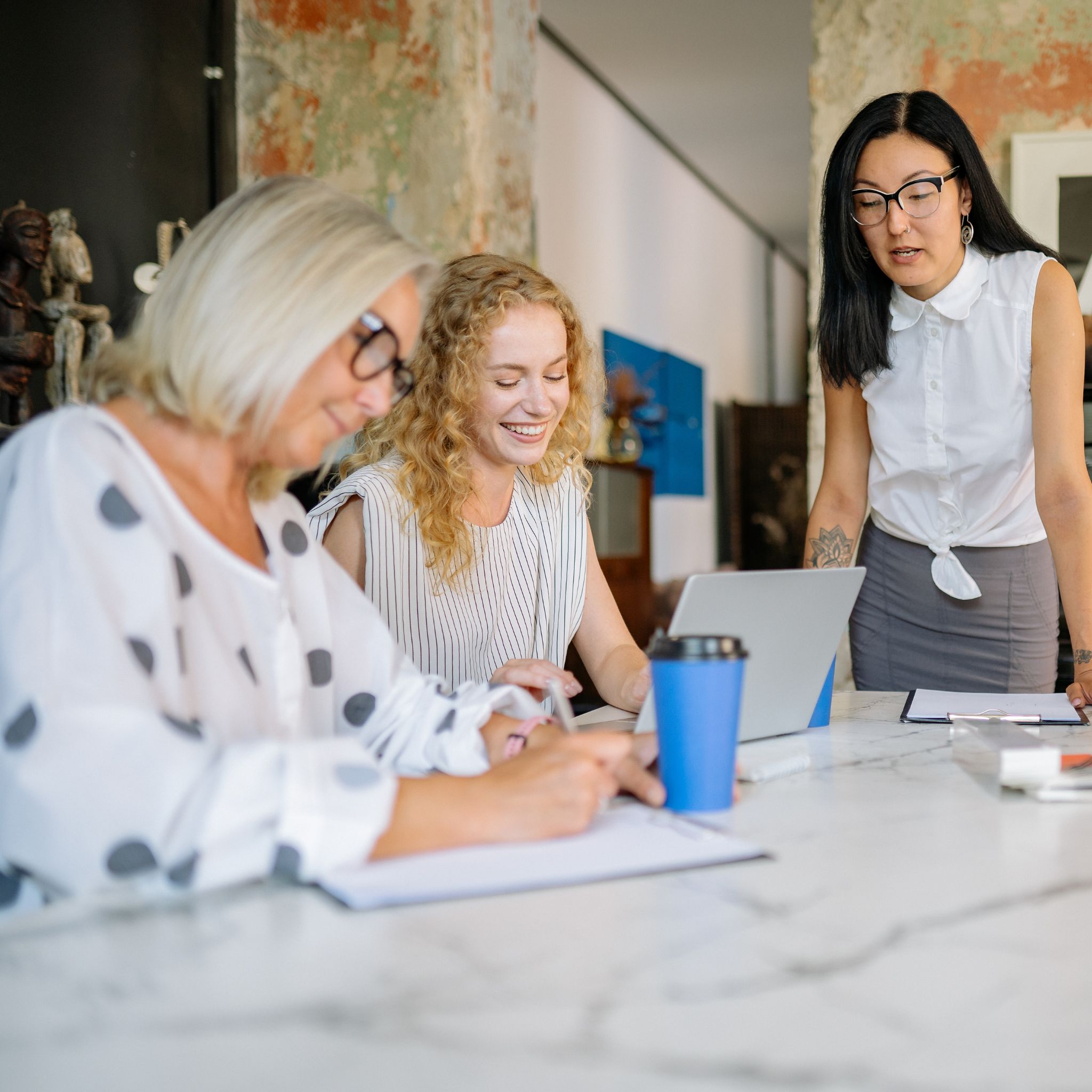 Three women around a table filling in a piece of paper. Two are sitting, one is standing, one has the pencil in hand