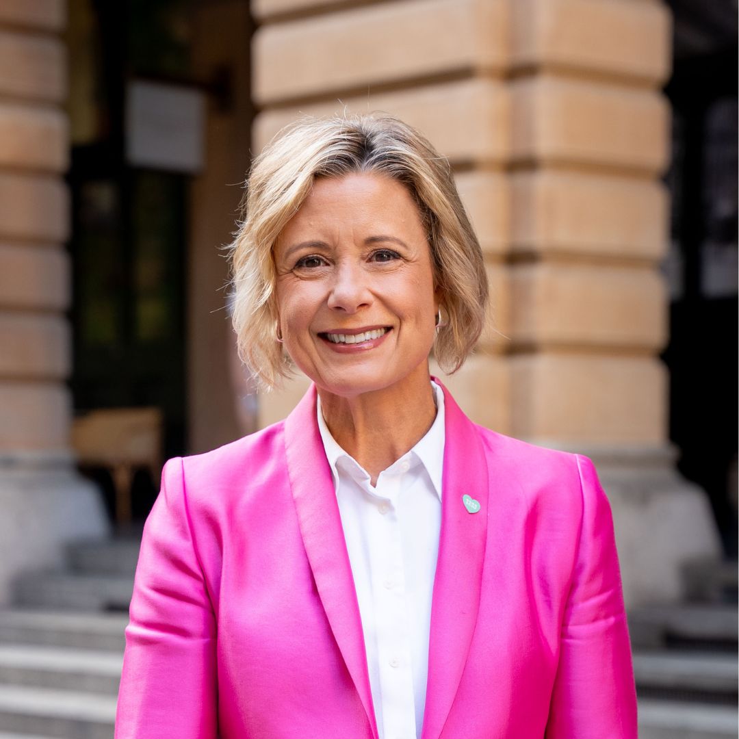 Headshot of Kristina Keneally who has short, blonde, wavy hair and wears a white button-down shirt with a bright pink jacket.