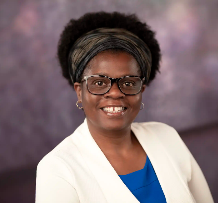 Headshot of Virginia Mapedzahama who has black, afro hair worn up with a dark headband, and wears black-rimmed glasses, a blue blouse and a white jacket. 