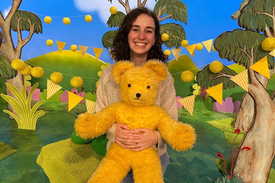 Photo of Belinda siting in front of a brightly coloured background holding a yellow teddy bear